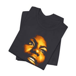 May include: A dark charcoal gray t-shirt featuring a high-contrast, golden-yellow graphic print of a person's face. The portrait focuses on the eyes, nose, and mouth, with deep shadows creating a dramatic effect against the dark fabric background.