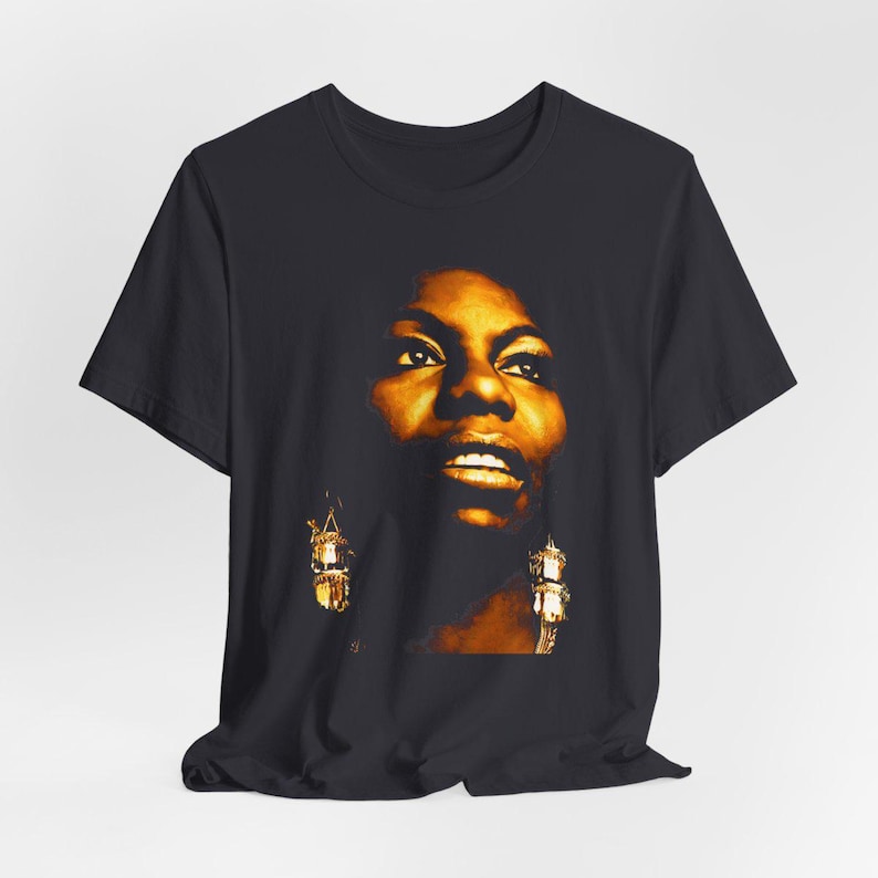 May include: A black short-sleeved t-shirt featuring a high-contrast, golden-toned graphic print of a person's face. The portrait highlights facial features with warm amber lighting and includes detailed depictions of dangling earrings.