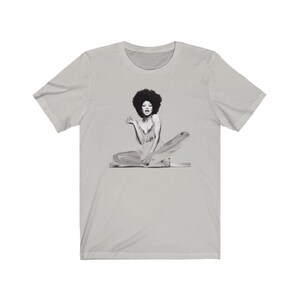 May include: A gray t-shirt with a black and white image of a woman with an afro sitting on a platform.