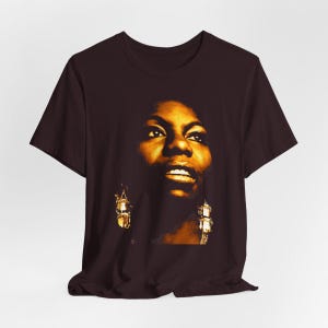 May include: A dark brown short-sleeved t-shirt featuring a large, high-contrast golden-yellow graphic print of a person's face. The portrait shows detailed facial features, including eyes, nose, and lips, with prominent dangling earrings visible.