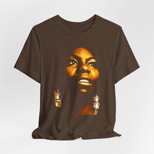 May include: A dark brown short-sleeved t-shirt featuring a large, centered graphic print of a person's face in warm golden and amber tones. The portrait shows detailed facial features, including eyes and lips, with dangling earrings visible.