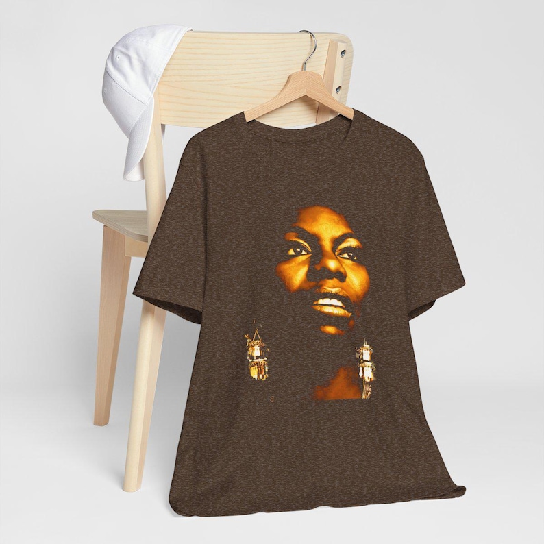 May include: A brown short-sleeve t-shirt featuring a graphic print of a person's face in warm golden and dark tones. The person has a focused expression and is wearing dangling earrings. The shirt is displayed on a wooden hanger.