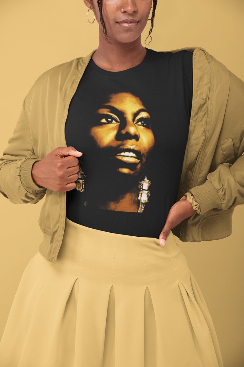May include: A black short-sleeved t-shirt featuring a high-contrast, golden-toned photographic portrait of a person with prominent earrings. The shirt is worn under a tan bomber jacket and paired with a tan pleated skirt.