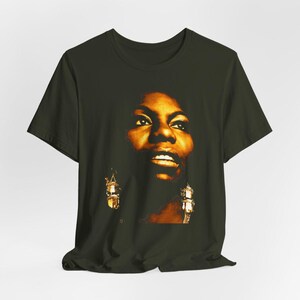 May include: A dark olive green short-sleeve t-shirt featuring a high-contrast, warm-toned graphic print of a person's face. The portrait is rendered in shades of gold, amber, and deep brown, highlighting facial features and dangling earrings.