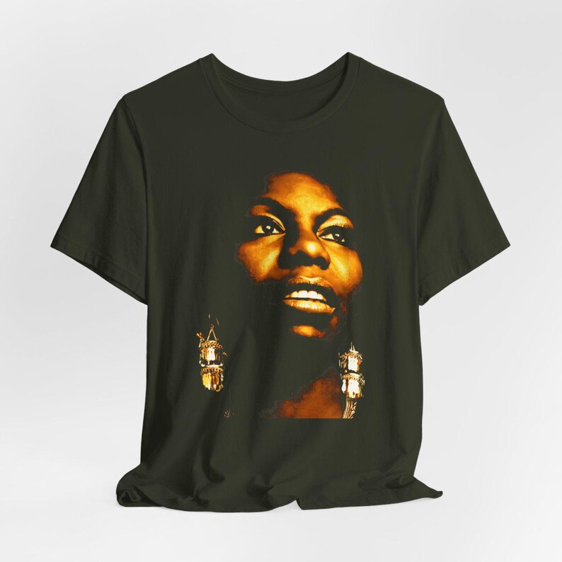 May include: A dark olive green short-sleeve t-shirt featuring a high-contrast, warm-toned graphic print of a person's face. The portrait is rendered in shades of gold, amber, and deep brown, highlighting facial features and dangling earrings.