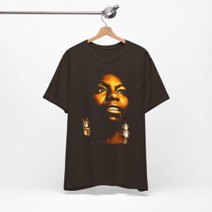 May include: A dark brown short-sleeve t-shirt featuring a high-contrast, warm-toned photographic print of a person's face on the front. The graphic highlights facial features with golden lighting and shows the person wearing dangling earrings.