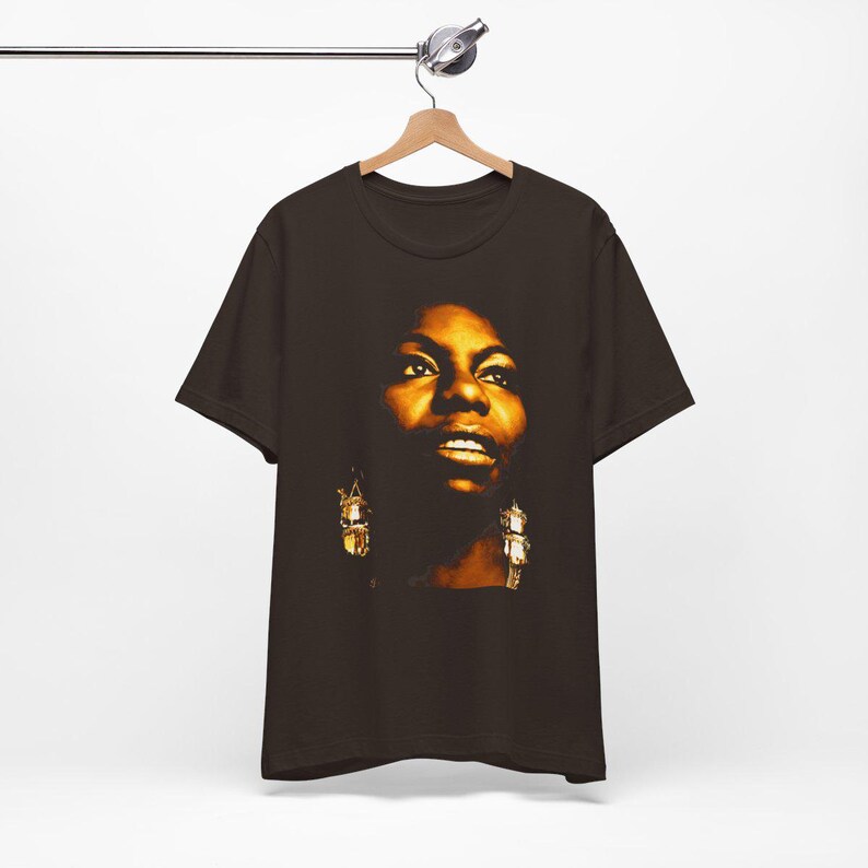 May include: A dark brown short-sleeve t-shirt featuring a high-contrast, warm-toned photographic print of a person's face on the front. The graphic highlights facial features with golden lighting and shows the person wearing dangling earrings.