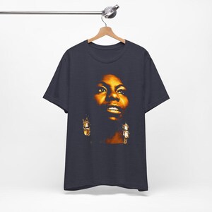 May include: A dark charcoal gray short-sleeve t-shirt featuring a high-contrast, warm-toned photographic print of a person's face centered on the chest. The portrait highlights facial features in shades of gold, amber, and deep brown shadows.