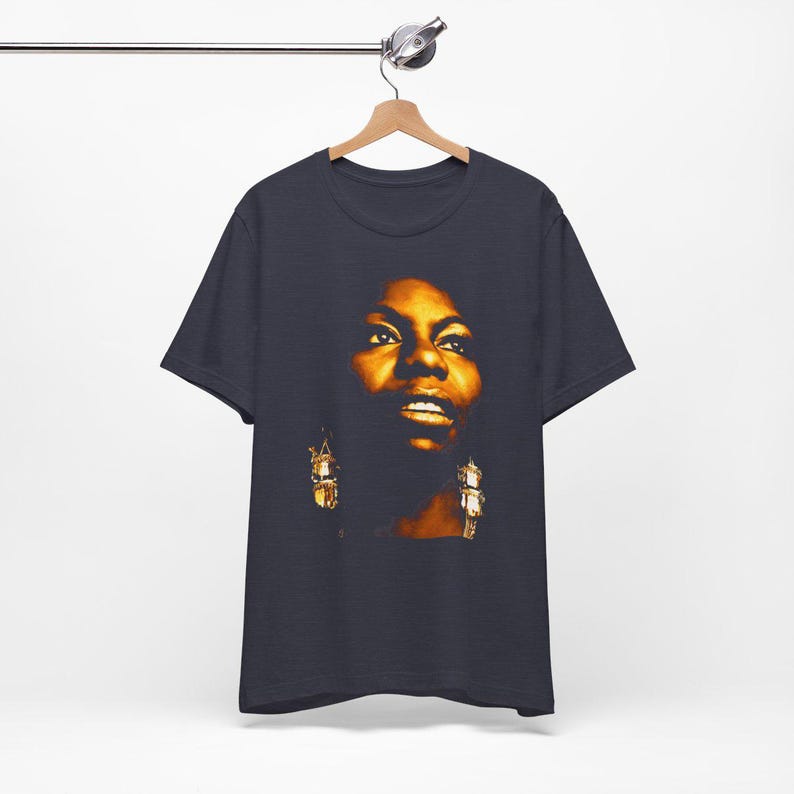 May include: A dark charcoal gray short-sleeve t-shirt featuring a high-contrast, warm-toned photographic print of a person's face centered on the chest. The portrait highlights facial features in shades of gold, amber, and deep brown shadows.