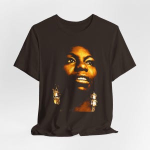 May include: A dark brown short-sleeved t-shirt featuring a high-contrast, golden-toned photographic print of a person's face. The graphic highlights facial features with deep shadows and bright highlights, including visible dangling earrings.