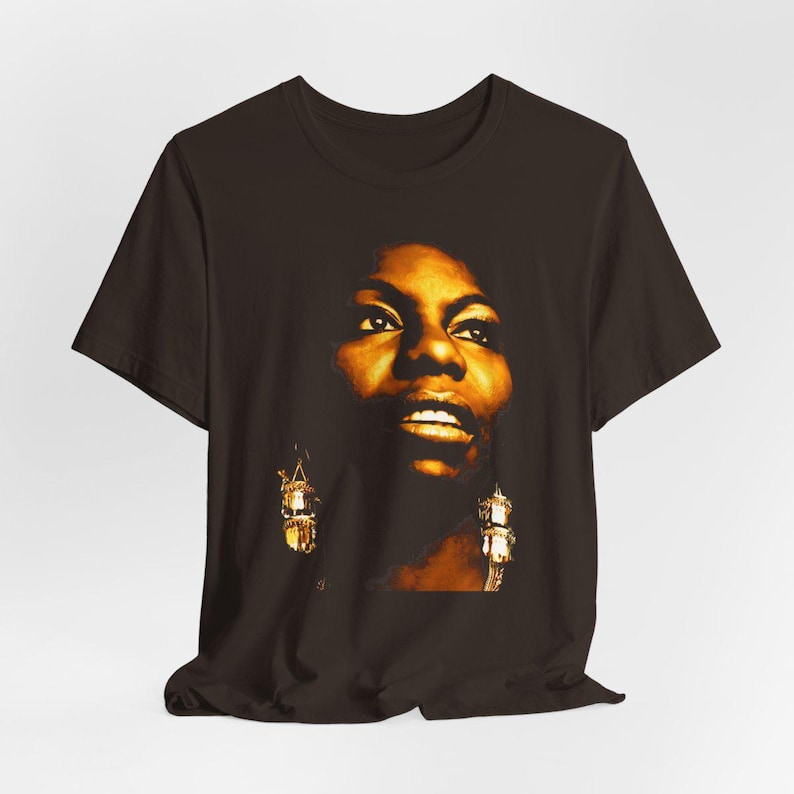 May include: A dark brown short-sleeved t-shirt featuring a high-contrast, golden-toned photographic print of a person's face. The graphic highlights facial features with deep shadows and bright highlights, including visible dangling earrings.