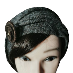 Pode incluir: Um chapéu cloche de lã cinza escuro com uma roseta decorativa marrom na lateral. O chapéu é estilizado em uma cabeça de manequim com cabelo escuro e batom vermelho.