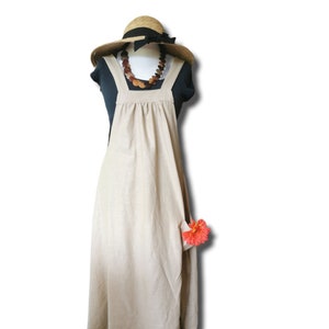 Puede incluir: Vestido delantal de lino beige con escote cuadrado y tirantes anchos. Un sombrero de paja con una cinta negra y un collar de cuentas de madera están encima. Una flor naranja brillante está sujeta al bolsillo. El vestido se exhibe en un maniquí.