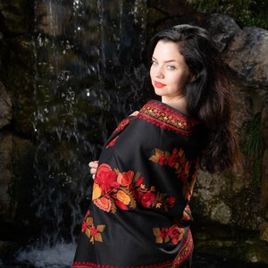 Op de afbeelding: Een vrouw draagt een zwarte sjaal met rode en oranje bloemenborduurwerk. De sjaal is over haar schouders gedrapeerd en ze staat voor een waterval.