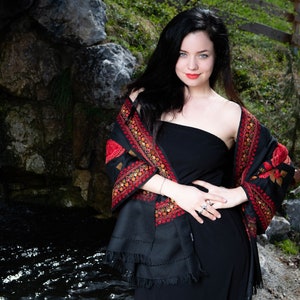 Op de afbeelding: Een vrouw draagt een zwarte strapless jurk met een zwarte sjaal met rode en gouden borduursels. De sjaal is over haar schouders gedrapeerd en vastgebonden in haar taille. Ze staat voor een rotsachtige achtergrond met een beek in de voorgrond.