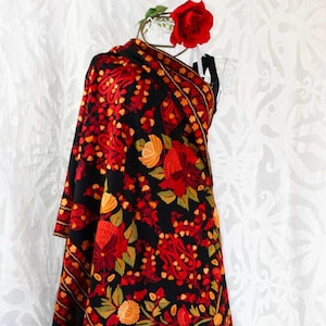 Puede incluir: Un chal negro con intrincados bordados florales rojos, naranjas y verdes. El chal tiene un borde con flecos y está drapeado sobre un maniquí.