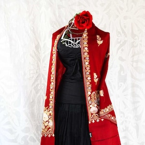 Puede incluir: Un chal rojo con bordados dorados y blancos. El chal tiene un borde de flecos y está drapeado sobre un maniquí negro.