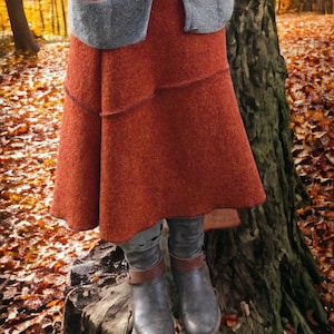 Könnte beinhalten: Ein rostfarbener Wollrock mit mehrschichtigem Design wird mit einer grauen Strickjacke kombiniert. Der Rock ist knielang und hat ein strukturiertes Aussehen. Graue Stiefel mit braunen Riemen vervollständigen das Outfit, vor dem Hintergrund von Herbstlaub.