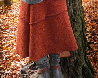 Può includere: Una gonna in lana color ruggine con un design a strati è abbinata a un cardigan grigio. La gonna è lunga fino al ginocchio e ha un aspetto strutturato. Stivali grigi con cinturini marroni completano l'outfit, sullo sfondo di un fogliame autunnale.