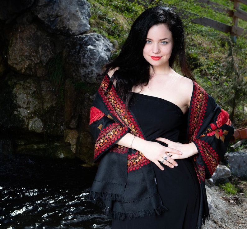 Op de afbeelding: Een vrouw draagt een zwarte strapless jurk met een zwarte sjaal met rode en gouden borduursels. De sjaal is over haar schouders gedrapeerd en vastgebonden in haar taille. Ze staat voor een rotsachtige achtergrond met een beek in de voorgrond.