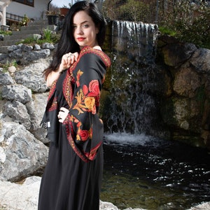 Op de afbeelding: Een vrouw in een zwarte jurk met een kleurrijke bloemenprint sjaal staat voor een kleine waterval. De sjaal heeft rode, oranje en gele bloemen op een zwarte achtergrond.