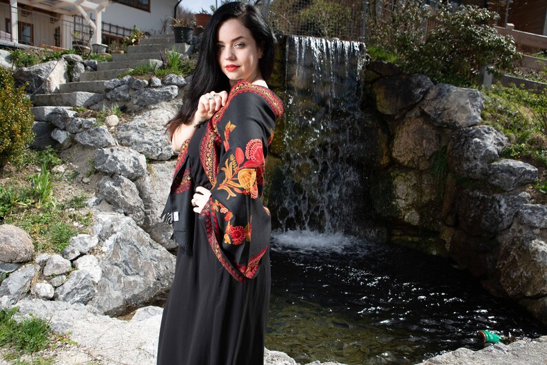 Op de afbeelding: Een vrouw in een zwarte jurk met een kleurrijke bloemenprint sjaal staat voor een kleine waterval. De sjaal heeft rode, oranje en gele bloemen op een zwarte achtergrond.