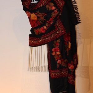 Op de afbeelding: Een zwarte sjaal met ingewikkelde bloemenborduurwerk in tinten rood, oranje en geel. De sjaal is gedrapeerd over een wit mannequin en heeft een franje rand.
