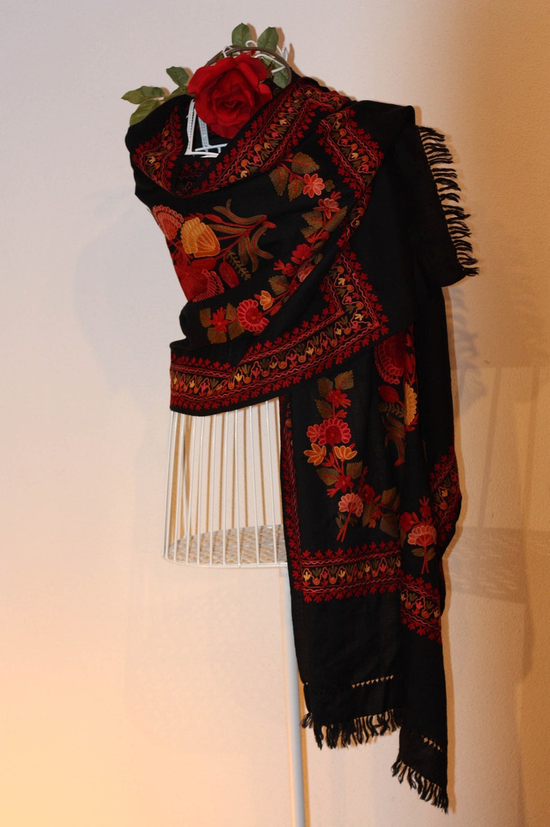 Op de afbeelding: Een zwarte sjaal met ingewikkelde bloemenborduurwerk in tinten rood, oranje en geel. De sjaal is gedrapeerd over een wit mannequin en heeft een franje rand.