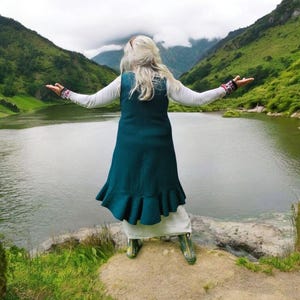 Puede incluir: Una persona está de pie con los brazos extendidos, frente a un lago y montañas. Lleva un chaleco color verde azulado con un dobladillo con volantes sobre un vestido blanco y botas verdes. La escena está ambientada al aire libre con un cielo nublado y verdes colinas.