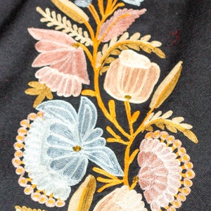 Op de afbeelding: Geborduurde bloemenontwerp op een zwarte stoffen achtergrond. Het ontwerp bevat een verscheidenheid aan bloemen in tinten roze, blauw en wit, met gouden en bruine stelen en bladeren.