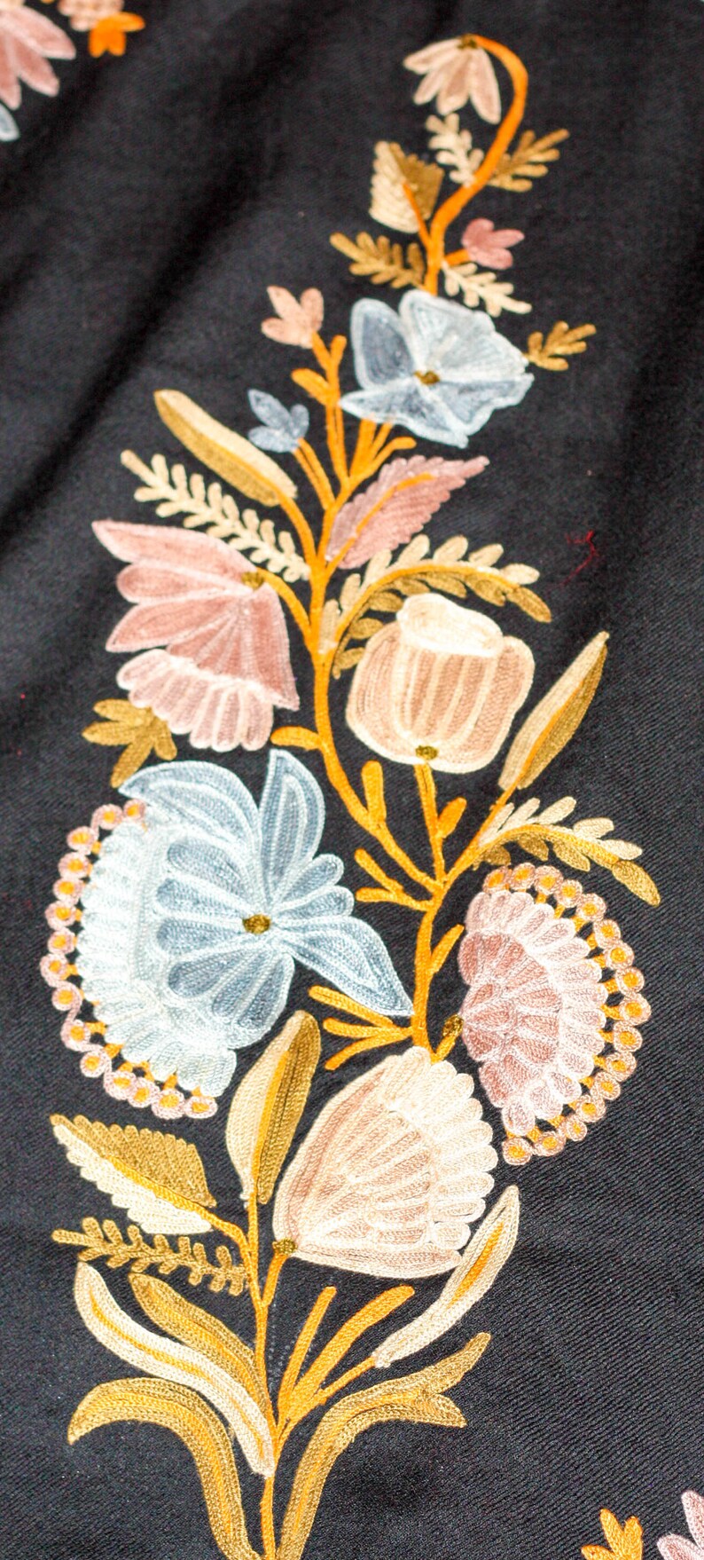 Op de afbeelding: Geborduurde bloemenontwerp op een zwarte stoffen achtergrond. Het ontwerp bevat een verscheidenheid aan bloemen in tinten roze, blauw en wit, met gouden en bruine stelen en bladeren.