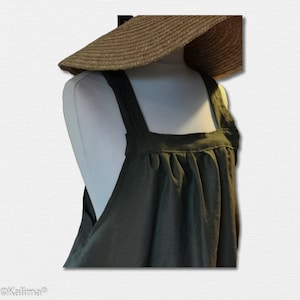 Puede incluir: Un delantal de lino verde oscuro con tirantes anchos y cintura fruncida. El delantal está en un maniquí blanco y un sombrero de paja marrón está en la cabeza del maniquí.