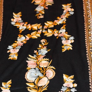 Op de afbeelding: Een zwarte sjaal met ingewikkelde bloemenborduurwerk in tinten roze, wit en goud. De sjaal heeft een decoratieve rand met een herhalend patroon.