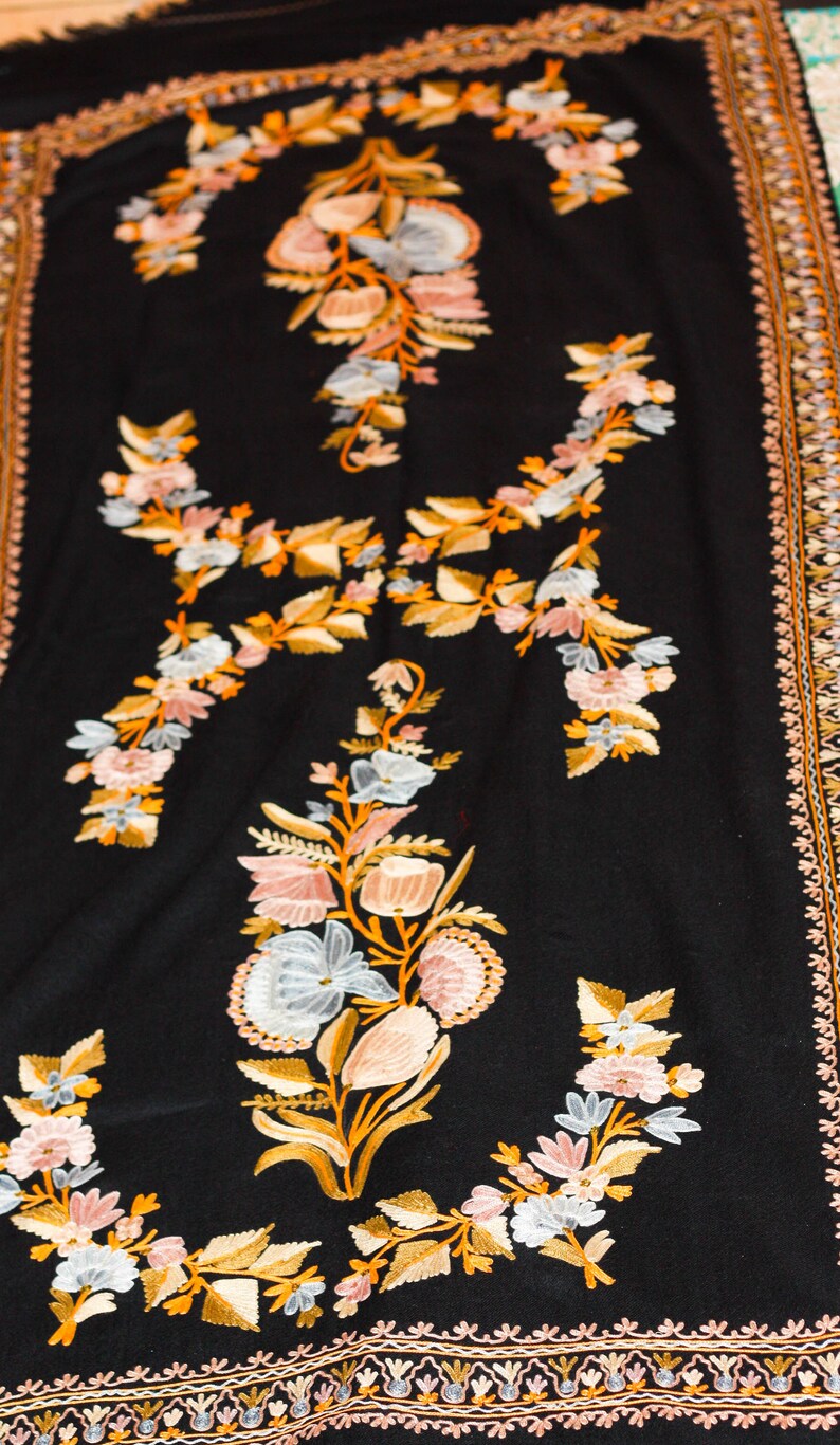 Op de afbeelding: Een zwarte sjaal met ingewikkelde bloemenborduurwerk in tinten roze, wit en goud. De sjaal heeft een decoratieve rand met een herhalend patroon.