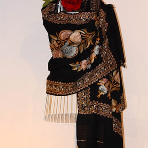 Op de afbeelding: Een zwarte sjaal met ingewikkelde bloemenborduurwerk in tinten goud, bruin en blauw. Een rode roos is bovenop de sjaal geplaatst.