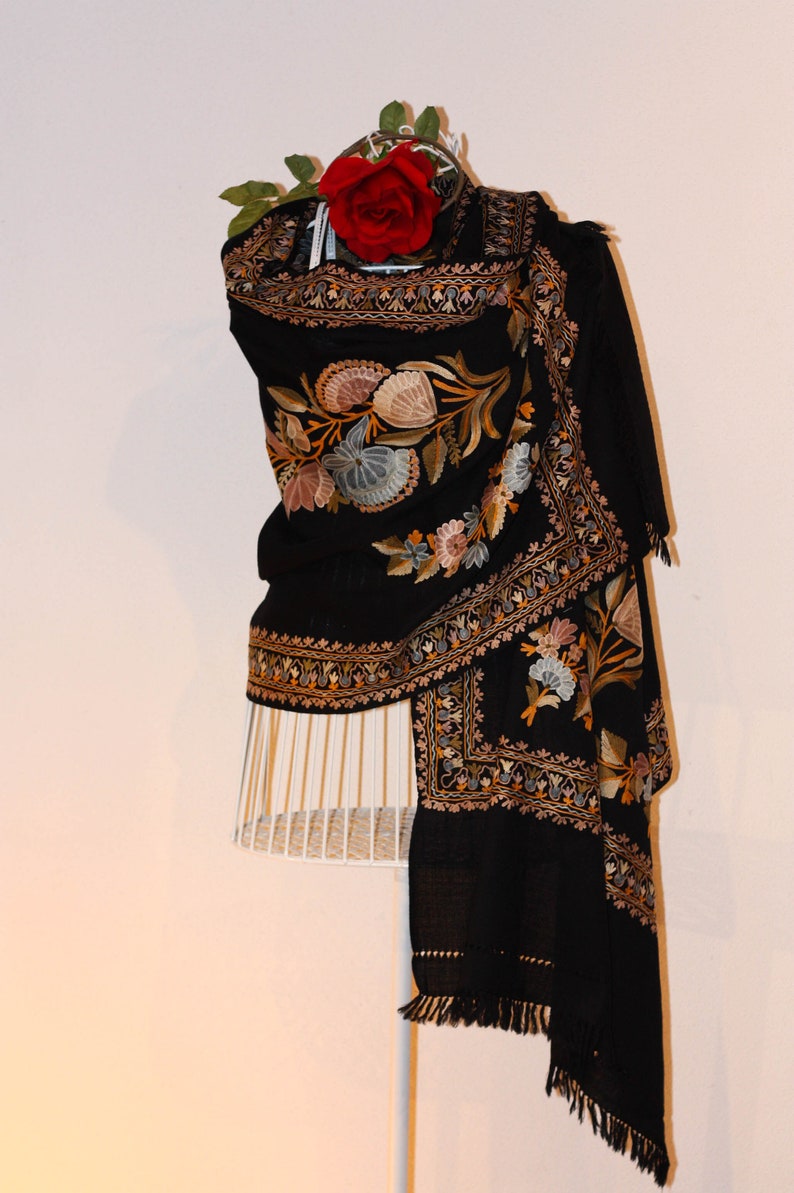Op de afbeelding: Een zwarte sjaal met ingewikkelde bloemenborduurwerk in tinten goud, bruin en blauw. Een rode roos is bovenop de sjaal geplaatst.