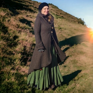 Op de afbeelding: Een vrouw in een bruine capuchonjas met een groene rok die op een grasachtige heuvel staat.