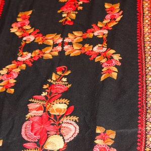 Op de afbeelding: Een zwarte sjaal met ingewikkelde borduursels van rode, oranje en roze bloemen. De sjaal heeft een decoratieve rand met een herhalend patroon.
