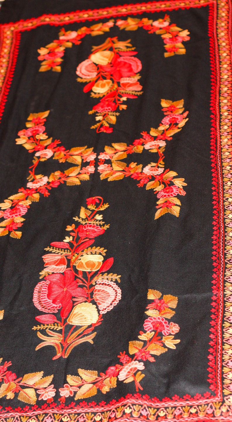 Op de afbeelding: Een zwarte sjaal met ingewikkelde borduursels van rode, oranje en roze bloemen. De sjaal heeft een decoratieve rand met een herhalend patroon.