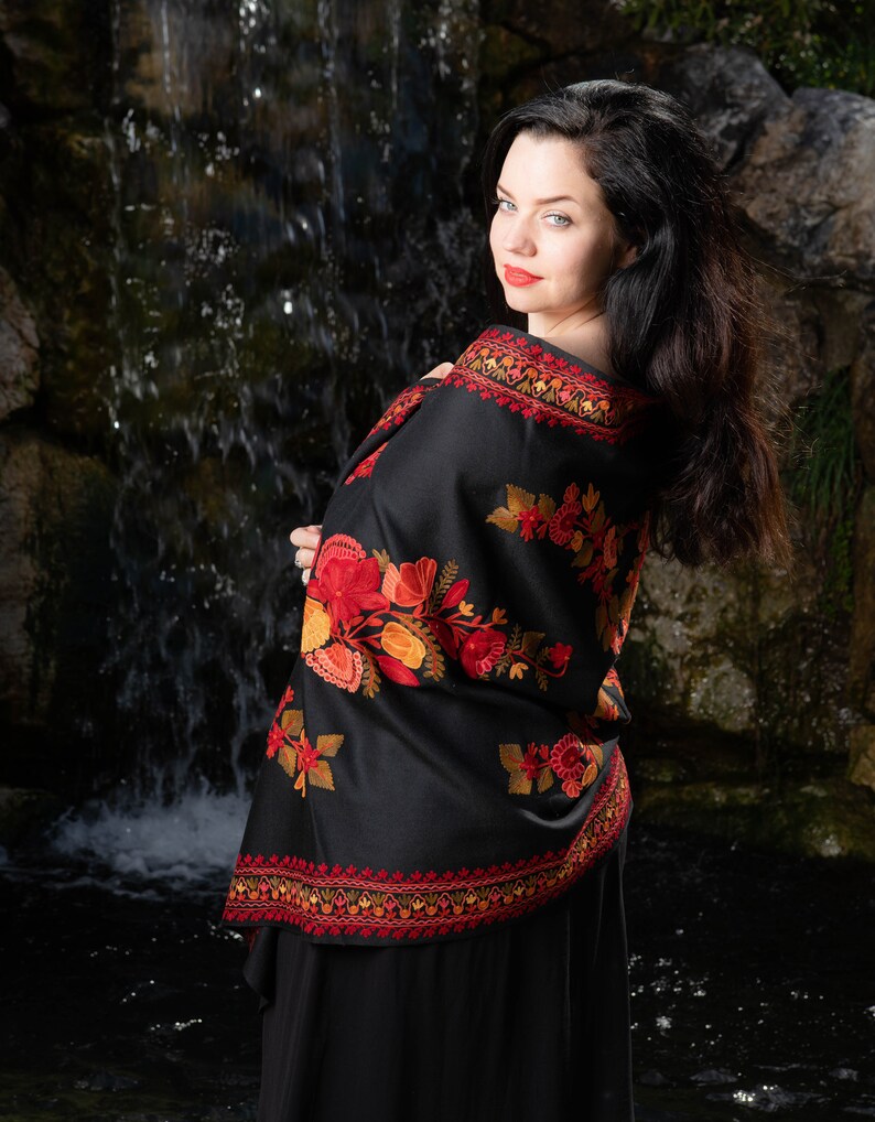 Op de afbeelding: Een vrouw draagt een zwarte sjaal met rode en oranje bloemenborduurwerk. De sjaal is over haar schouders gedrapeerd en ze staat voor een waterval.