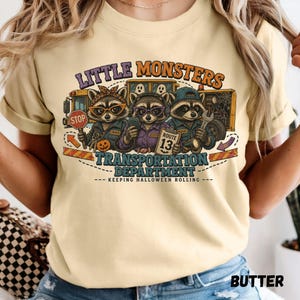 May include: A pale yellow t-shirt featuring a graphic of three raccoons dressed as transportation workers in front of a school bus. The text reads LITTLE MONSTERS TRANSPORTATION DEPARTMENT KEEPING HALLOWEEN ROLLING and ROUTE 13.