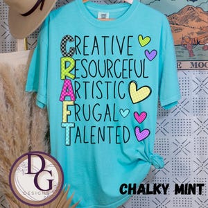 以下が含まれることがあります： カラフルな模様の文字で「CRAFT」と書かれたターコイズ色のTシャツ。「CREATIVE, RESOURCEFUL, ARTISTIC, FRUGAL, TALENTED」の文字が「CRAFT」の上下に書かれています。Tシャツの下部には「CHALKY MINT」の文字も書かれています。