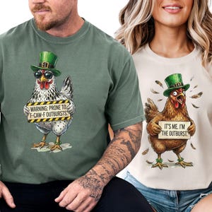 May include: Two t-shirts with chicken graphics. One is sage green with a chicken wearing sunglasses and a green hat, holding a sign that reads "WARNING: PRONE TO F-CAW-F OUTBURSTS." The other is cream-colored with a chicken wearing a green hat and holding a sign that reads "IT'S ME. I'M THE OUTBURST."