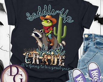Puede incluir: Camiseta negra con un dibujo de una rana con sombrero de vaquero montada en un mapache, con un cactus. El texto dice "Saddle Up First Grade" y "It's going to be a great year."