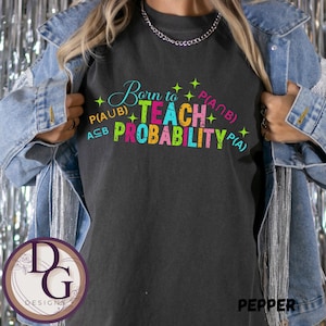 May include: Dark gray t-shirt with the text "Born to Teach Probability" in colorful letters. The shirt also features mathematical symbols and the brand name "Pepper".
