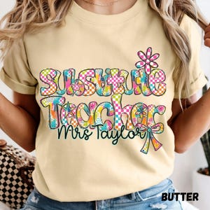 Puede incluir: Una camiseta de color amarillo claro con el texto "Substitute Teacher" y "Mrs Taylor" en una fuente colorida, a cuadros y con salpicaduras de pintura. El diseño incluye un pequeño gráfico de flor sobre el texto y un lazo decorativo.