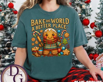 Könnte beinhalten: Ein teal-farbenes T-Shirt mit dem Text "BAKE THE WORLD A BETTER PLACE". Die Grafik zeigt einen Lebkuchenmann mit Nudelholz, umgeben von Weihnachtsplätzchen und -ornamenten. Teil der "BLUE SPRUCE" Kollektion.