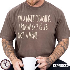 Könnte beinhalten: Espressofarbenes T-Shirt mit dem weißen Text: "I'M A MATH TEACHER. I KNOW 6+7 IS 13 NOT A MEME." Das Shirt hat einen Rundhalsausschnitt. Eine Person hält eine weiße Tasse.