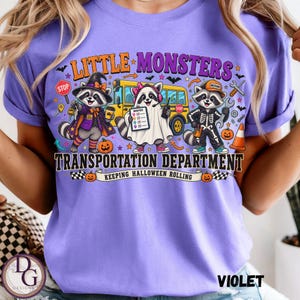 May include: A light purple t-shirt featuring a graphic of three raccoons dressed in Halloween costumes in front of a yellow school bus. The text reads LITTLE MONSTERS TRANSPORTATION DEPARTMENT KEEPING HALLOWEEN ROLLING with various icons.