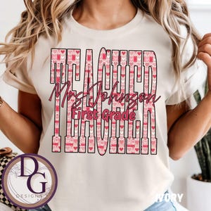 Peut inclure: T-shirt ivoire avec le mot "TEACHER" en grandes lettres roses remplies d'un motif de cœurs. En dessous, en écriture cursive, on lit "Mrs. Johnson First Grade". Un logo DG Designs est en bas à gauche.